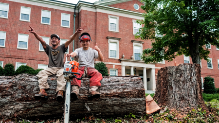 Two male workers smiling and posing for a photo sitting atop a chopped down tree in front a brick building.